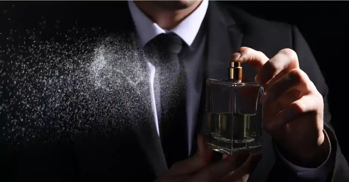 A man in black suit is holding a perfume in his hand while a mist is being sprayed from the bottle